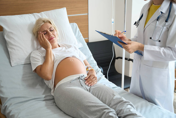 Teilweise weibliche Ärztin schreibt in Klemmbrett über erwachsene kaukasische schwangere Frau mit Kopfschmerzen auf medizinischem Bett, während sie in der Geburtsklinik untersucht wird - Foto, Bild
