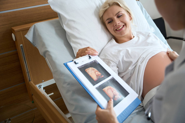 Sonriente mujer embarazada caucásica adulta en la cama médica mirando la foto de ultrasonido de su futuro bebé en la mano de la doctora recortada en el hospital de maternidad - Foto, Imagen