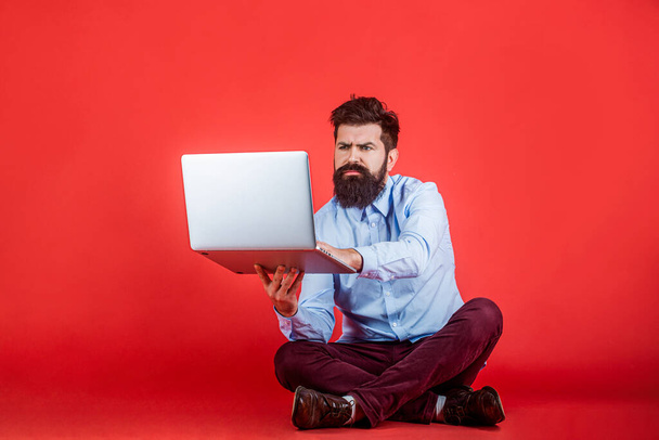 Ciddi sakallı işçi dizüstü bilgisayarı. Erkek işçi bilgisayarı. Sakallı adam kırmızı arka planda dizüstü bilgisayarla yerde oturuyor. Dizüstü bilgisayarı tutuyor. Genç işadamı bilgisayarını kullanıyor.. - Fotoğraf, Görsel
