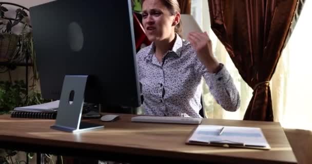 Irritada mujer enojada sentada en la mesa al lado de la computadora y jura por el monitor roto. Malware y mal funcionamiento y reparación de computadoras concepto - Metraje, vídeo