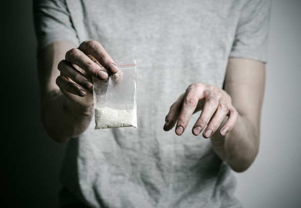 The fight against drugs and drug addiction topic: addict holding package of cocaine in a gray T-shirt on a dark background in the studio - Fotografie, Obrázek
