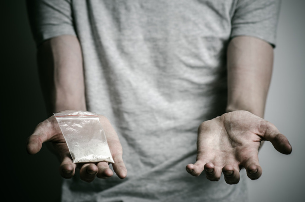 The fight against drugs and drug addiction topic: addict holding package of cocaine in a gray T-shirt on a dark background in the studio - Photo, Image