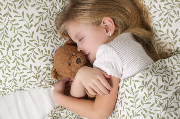 Tatlı küçük kız yatakta uyuyor ve oyuncak ayısına sarılıyor. - Fotoğraf, Görsel