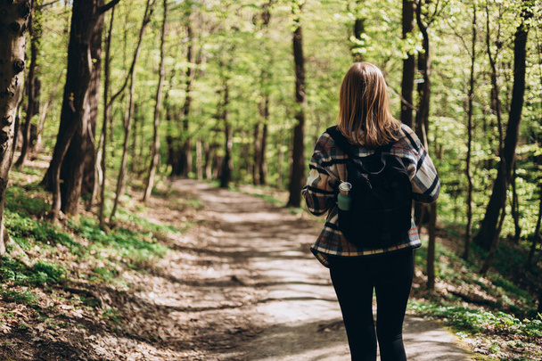 Rear view of young woman with casual outfit, wears a black backpack walking in the forest. Spring time. 30s Women in sport clothes hiking in forest - Photo, Image