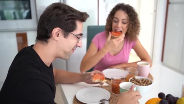 Reaaliaikainen onnellinen aikuinen pari, keskustellessaan ja katsoessaan televisiota istuessaan ruokapöydässä syöden leipää hedelmähillolla ja nauraen pienen inkiväärin näyttämisestä päivänvalossa - Materiaali, video