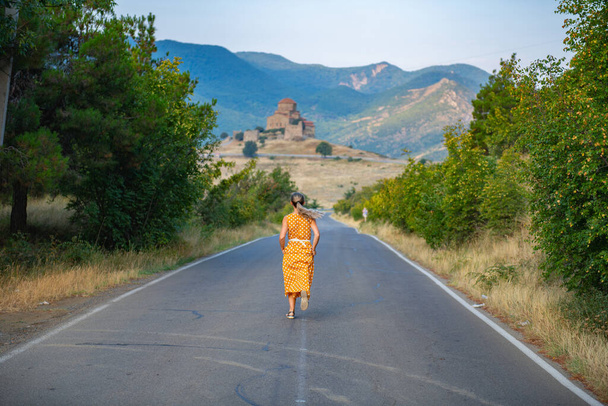Een eenzaam meisje in een opvallende oranje kledij loopt rustig langs een charmante weg die zich uitstrekt naar een serene kerk en een majestueuze bergketen in de verte. - Foto, afbeelding