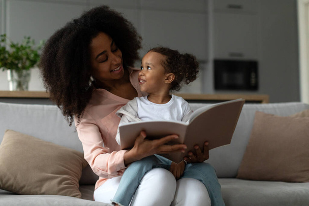 Rakastava musta äiti lukee kirjaa pienen tyttärensä kanssa, istuu yhdessä sohvalla kotona, viettää aikaa yhdessä olohuoneessa - Valokuva, kuva