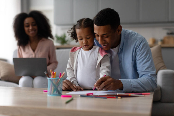 Aimer père noir enseigner le dessin de petite fille pendant que la mère travaille sur ordinateur portable sur fond, assis sur le canapé à la maison - Photo, image