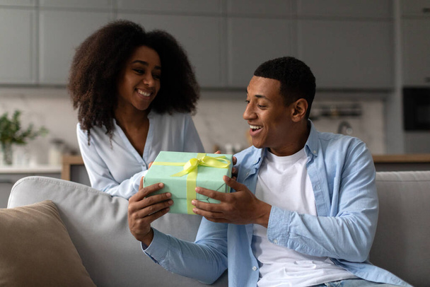 ハッピーアフリカ系アメリカ人女性はギフトボックスを与え,夫に驚きを与え,自宅でソファに座って笑顔で - 写真・画像