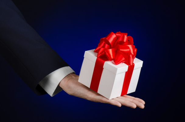 Il tema delle celebrazioni e dei regali: un uomo in abito nero con in mano un regalo esclusivo avvolto in una scatola bianca con nastro rosso, bellissimo e costoso regalo su uno sfondo blu scuro in studio isolato
 - Foto, immagini