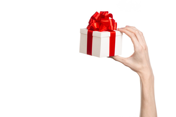 Het thema van vieringen en giften: hand met een geschenk verpakt in witte doos met rood lint en boog, het mooiste geschenk geïsoleerd op een witte achtergrond in studio - Foto, afbeelding
