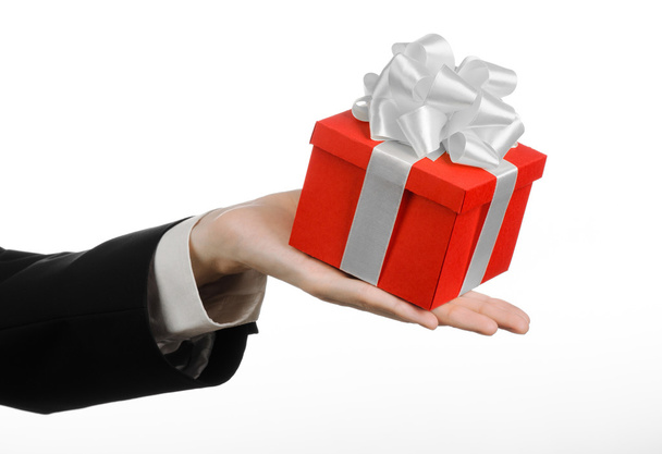Το θέμα της γιορτές και δώρα: ένα άτομο σε ένα μαύρο κοστούμι κρατώντας ένα αποκλειστικό δώρο τυλιγμένο σε κόκκινο κουτί με λευκή κορδέλα και φιόγκο, το πιο όμορφο δώρο που απομονώνονται σε λευκό φόντο σε στούντιο - Φωτογραφία, εικόνα