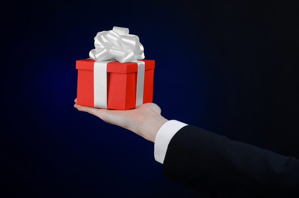 Il tema delle celebrazioni e dei regali: un uomo in abito nero con in mano un regalo esclusivo avvolto in scatola rossa con nastro bianco, bellissimo e costoso regalo su uno sfondo blu scuro in studio isolato
 - Foto, immagini