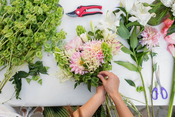Blick über die Hände der Floristen, die sorgfältig einen Strauß mit rosa Dahlien, Grün und anderen Blumen auf dem Tisch arrangieren - Foto, Bild
