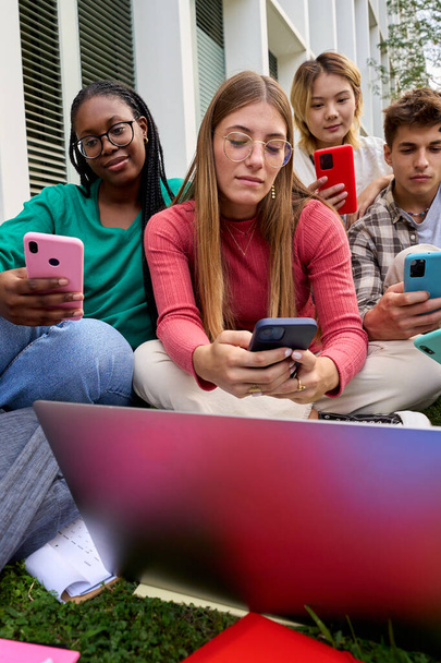 Κάθετη ομάδα νεαρών πολυφυλετικών φοιτητών, βυθισμένοι στο κινητό τους κινητό τηλέφωνο, εθισμένοι στην τεχνολογία φίλοι έξω από την πανεπιστημιούπολη. Gen z άτομα σοβαρή και συμπυκνωμένη χρήση smartphone - Φωτογραφία, εικόνα