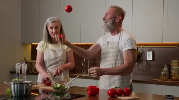 Valkoihoinen onnellinen perhe eläkkeellä keski-ikäinen pari rakastunut nainen ruoanlaitto aamiainen keittiössä hauska vanha mies jongleeraa tomaatteja tuoreita vihanneksia kypsä isoisä ja isoäiti hauskaa nauraa kotona - Materiaali, video