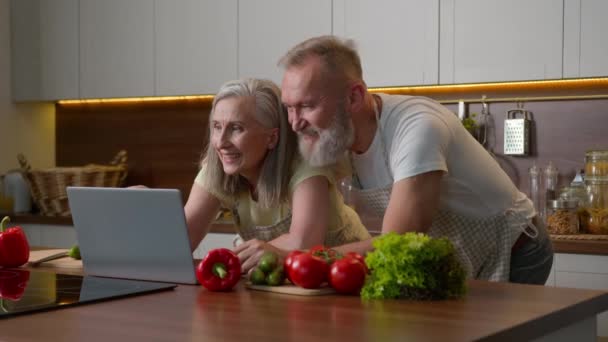Vanhemmat valkoihoinen pari keski-ikäiset puolisot perhe käyttää kannettavaa tietokonetta kotona keittiössä valitsemalla ruoka toimitus tuoreita vihanneksia verkossa ostoksia luonnontuotteita tilata eläkkeellä nainen ja mies nauraa hauskaa - Materiaali, video