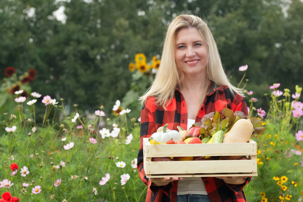 Uma mulher branca sorrindo está segurando uma caixa de madeira com legumes caseiros frescos variados (tomate, batata, pimenta, alface, abobrinha, abóbora, cebola) no jardim, florescendo flores silvestres no fundo. - Foto, Imagem