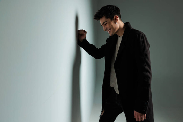 сексуальний щасливий чоловік в темному пальто торкаючись стіни, сміючись і дивлячись вниз, позуючи холодним способом перед сірим тлом  - Фото, зображення