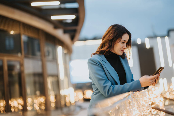 Una mujer de negocios en una chaqueta azul comprueba su teléfono en medio del resplandor de las luces de la ciudad durante una noche, retratando una sensación de conectividad y estilo de vida urbano. - Foto, imagen