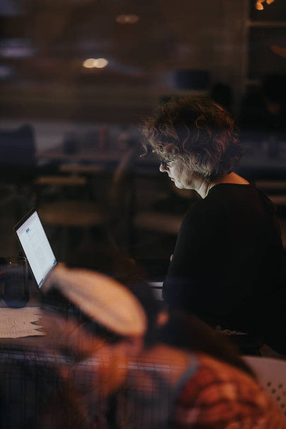 Profiel van een geconcentreerde vrouw die op een laptop werkt in een zwak verlicht kantoor. Een gevoel van toewijding en late nacht productiviteit komt voort uit de scène. - Foto, afbeelding