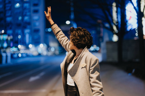 Uma mulher adulta sinaliza para um táxi em uma movimentada rua da cidade à noite, com efeito de luz ambiente criando uma vibração urbana. - Foto, Imagem