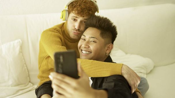 gay pareja abrazando y sonriendo mientras tomando selfie juntos sentado en el sofá - Foto, imagen