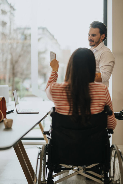 Discussão de negócios cuidadosa em um escritório moderno, com foco na inclusão, com uma mulher em uma cadeira de rodas segurando uma nota. - Foto, Imagem