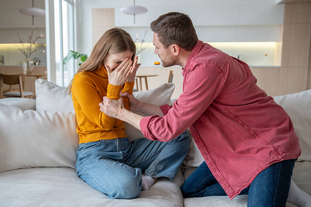Aggressive stressed tyrant man husband shouts grabs wife by arms. Unhappy scared woman crying closing face by hands sitting at home. Domestic physical, emotional violence, family quarrel, gaslighting. - Photo, Image