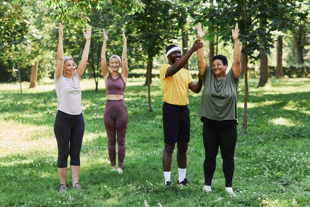 Idősek változatos csoportja dolgozik a szabadban a zöld parkban az edzővel és boldogan mosolyog. - Fotó, kép
