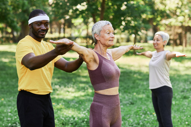 Retrato de cintura hacia arriba del entrenador sonriente que ayuda a la mujer mayor a hacer ejercicio al aire libre en el parque verde - Foto, Imagen