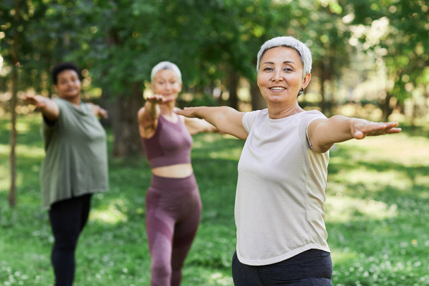 Waist up portrait of smiling senior woman enjoying sports workout outdoors and looking at camera while doing stretching exercise, copy space - Photo, Image