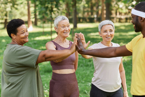 Группа активных женщин старшего возраста "дай пять" с тренером после занятий на открытом воздухе в парке - Фото, изображение