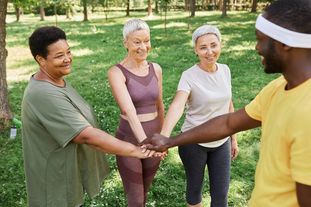 Groupe de femmes âgées actives empilant les mains avec un entraîneur après avoir apprécié l'entraînement en plein air dans le parc - Photo, image