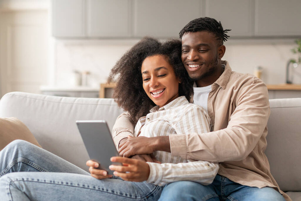 Šťastný černý mladý pár, který zkoumá obsah digitálního tabletu v interiéru, používá počítač a užívá si společně čas, odpočívá na gauči v moderním obývacím pokoji doma. Manželé sledující videa online - Fotografie, Obrázek