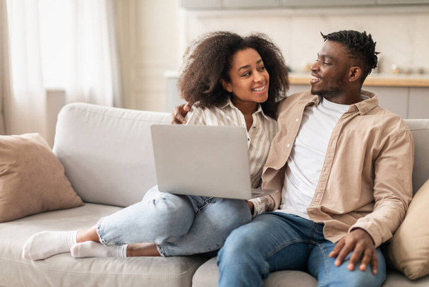Afro-americanos milenares cônjuges gostando de assistir vídeo on-line em seu PC laptop, relaxando no sofá no conforto da sala de estar em casa interior. Tecnologia on-line, lazer computador familiar - Foto, Imagem
