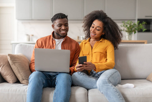 Great Online Offer. Happy Black Young Couple Reading Message On Smartphone And Using Laptop Computer, Sitting Together And Recommending Application Or Website On Sofa At Home - Photo, Image