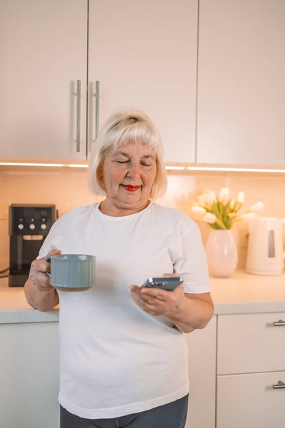 キッチンで自宅でリラックスしながら,新鮮な朝のコーヒーとテキストメッセージを飲む中年女性 - 写真・画像