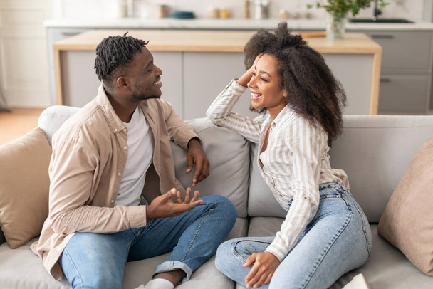 幸せな結婚したアフリカ系アメリカ人のカップルは,週末に家でソファーで座っています. 居心地の良いリビングルームで互いに笑顔のミレニアルブラックスパイ - 写真・画像