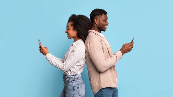 Pareja de jóvenes negros absortos de pie espalda con espalda, cada uno absorbido en su propio teléfono inteligente, destacando la interacción digital moderna sobre fondo azul - Foto, imagen