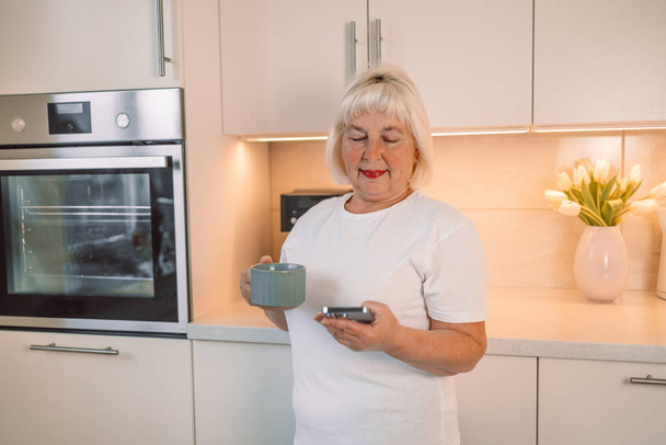 Middle aged woman drinking fresh morning coffee and text messaging while relaxing at home in her kitchen - Photo, Image