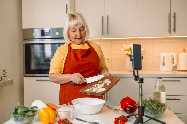 Caucasian female food blogger working on a new video and explaining how to cook a dish. Hands of senior 60s female chopping fresh onion on wooden board while preparing italian pasta with vegetables - Photo, Image