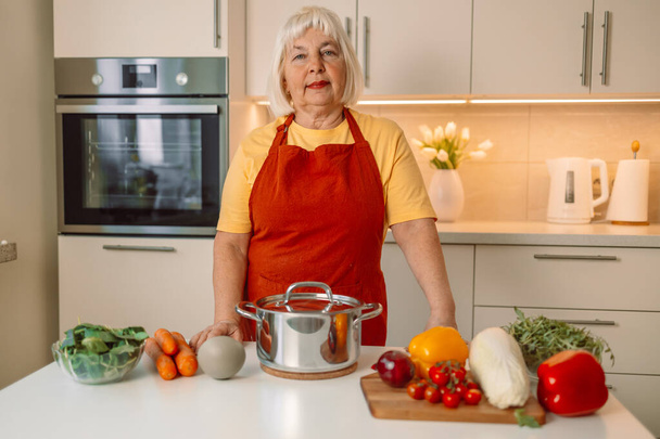 Krásná šťastná seniorka vaří v kuchyni a testuje polévku s čerstvou zeleninou z pánve na sporáku. Kvalitní fotografie - Fotografie, Obrázek