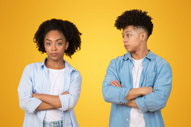 Sceptische jonge Afro-Amerikaanse vrouw en man met gekruiste armen, die elkaar een twijfelachtige blik geven, die wantrouwen of onenigheid symboliseren, tegen een levendige gele achtergrond - Foto, afbeelding