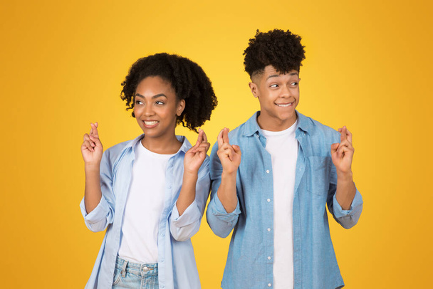 Оптимистичные счастливые афроамериканская молодая женщина и мужчина со скрещенными пальцами, обнадеживающие выражения и случайный стиль, стоя на желтом фоне, желая удачи - Фото, изображение
