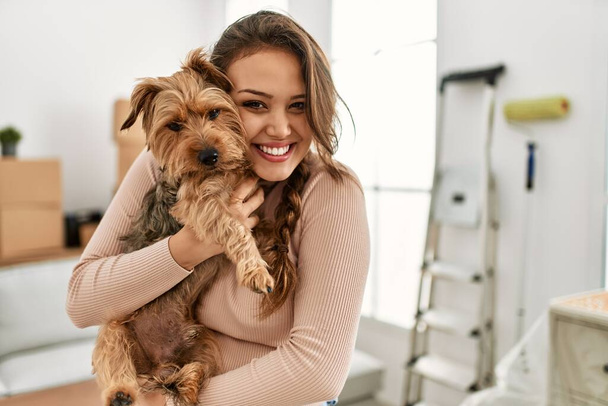 Joven hermosa mujer hispana sonriendo seguro abrazando perro en nuevo hogar - Foto, Imagen