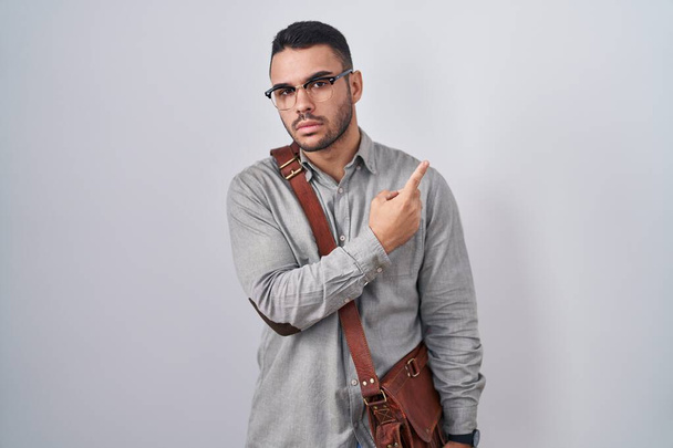 Young hispanic man wearing suitcase pointing with hand finger to the side showing advertisement, serious and calm face  - Photo, Image