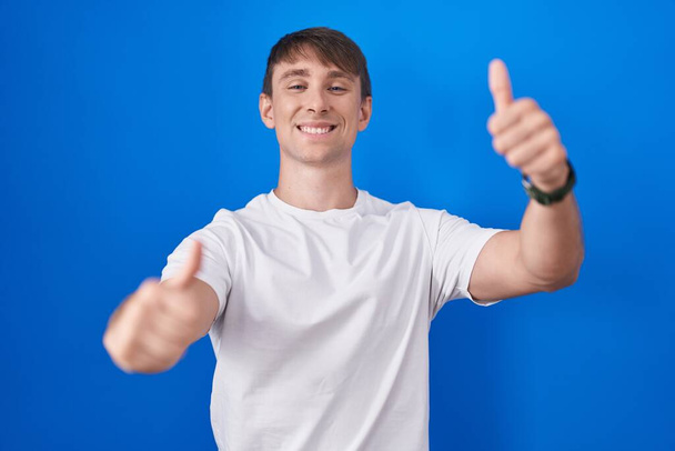 Blanke blonde man die over een blauwe achtergrond staat en positief gebaar maakt met de hand, glimlacht en blij voor succes. winnaar gebaar.  - Foto, afbeelding