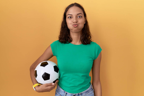 Молода іспаномовна жінка тримає м'яч набиваючи щоки зі смішним обличчям. рот надутий повітрям, божевільний вираз.  - Фото, зображення