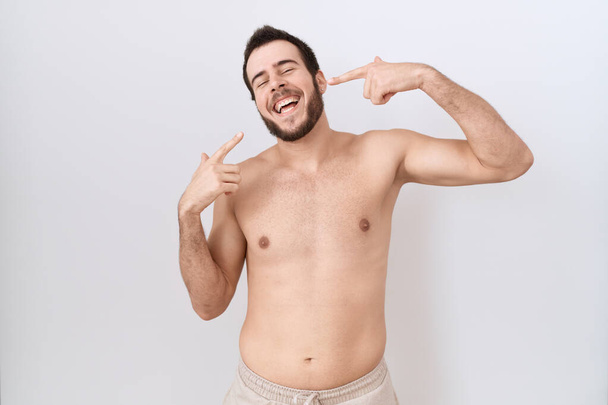 Jonge Spaanse man staat shirtloos over witte achtergrond glimlachend vrolijk tonen en wijzen met vingers tanden en mond. tandheelkundige gezondheidszorg.  - Foto, afbeelding